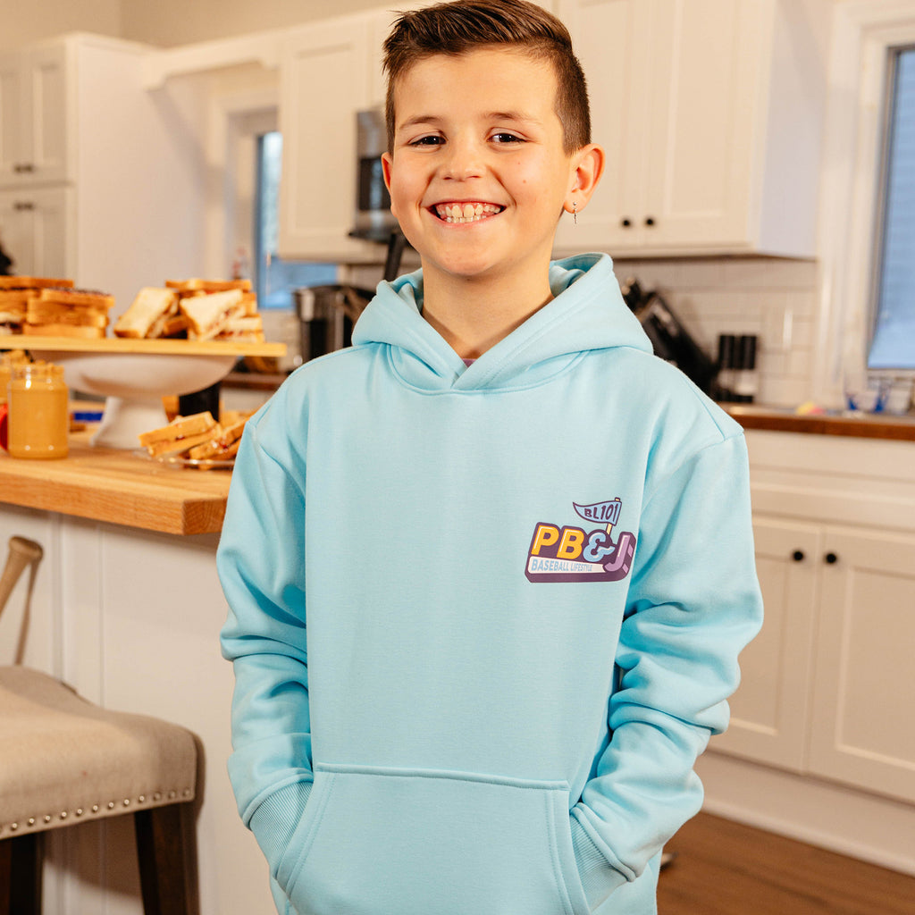 Young boy smiling in a kitchen wearing the BL101 youth PB&J Baseball Lifestyle light blue hoodie, showing the chest logo.