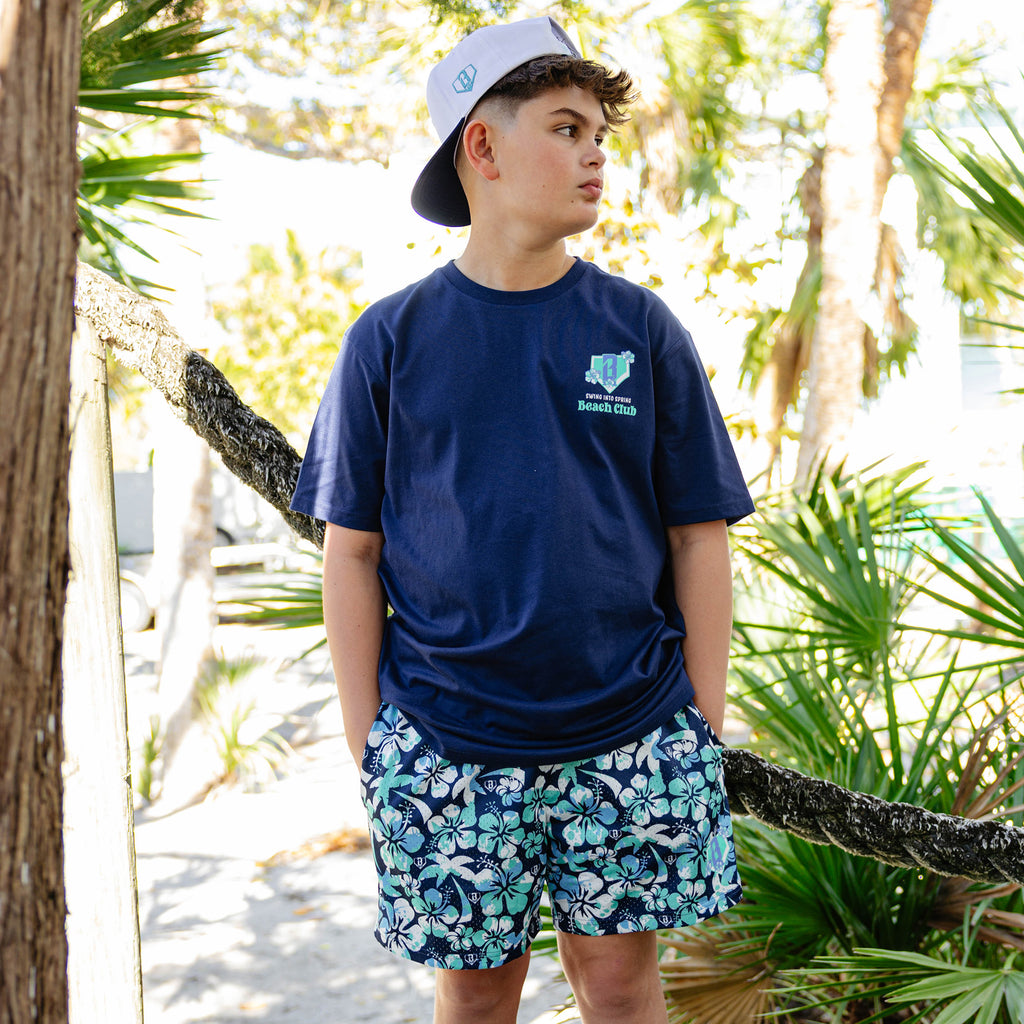 Person wearing a navy blue t-shirt with floral BL101 logo and floral shorts standing in a tropical setting.
