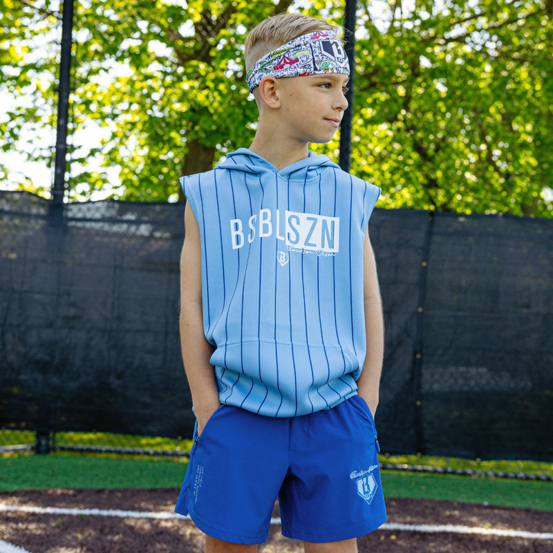 Youth player on a baseball field wearing the blue Jackson Olson x BL101 Pro Series Shorts V2 paired with a matching light blue pinstripe sleeveless hoodie and Jackson Olson graphic headband