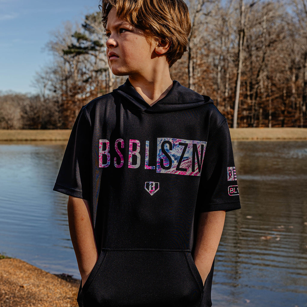 Child wearing a black hoodie with colorful 'BSBL SZN' by a body of water