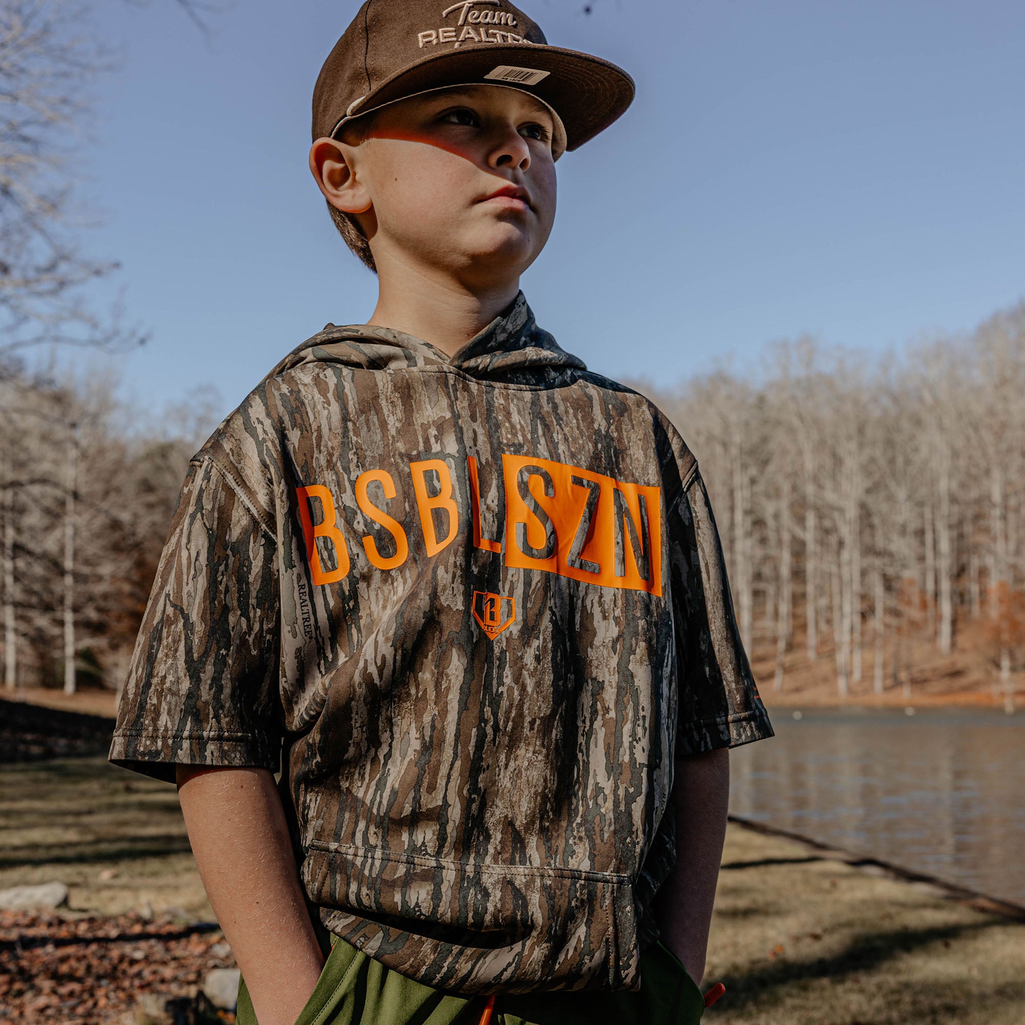 Child wearing a camouflage hoodie with 'BSBLSZN' text, standing by a body of water with trees in the background.