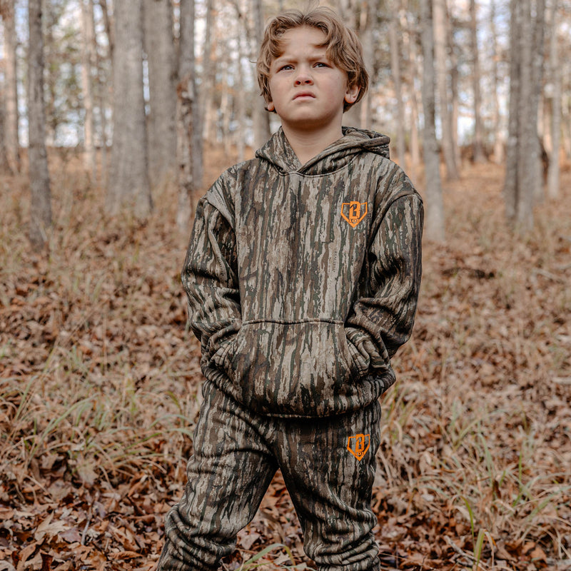 Child wearing a camouflage hoodie and pants with orange BL101 logo standing in a forest.