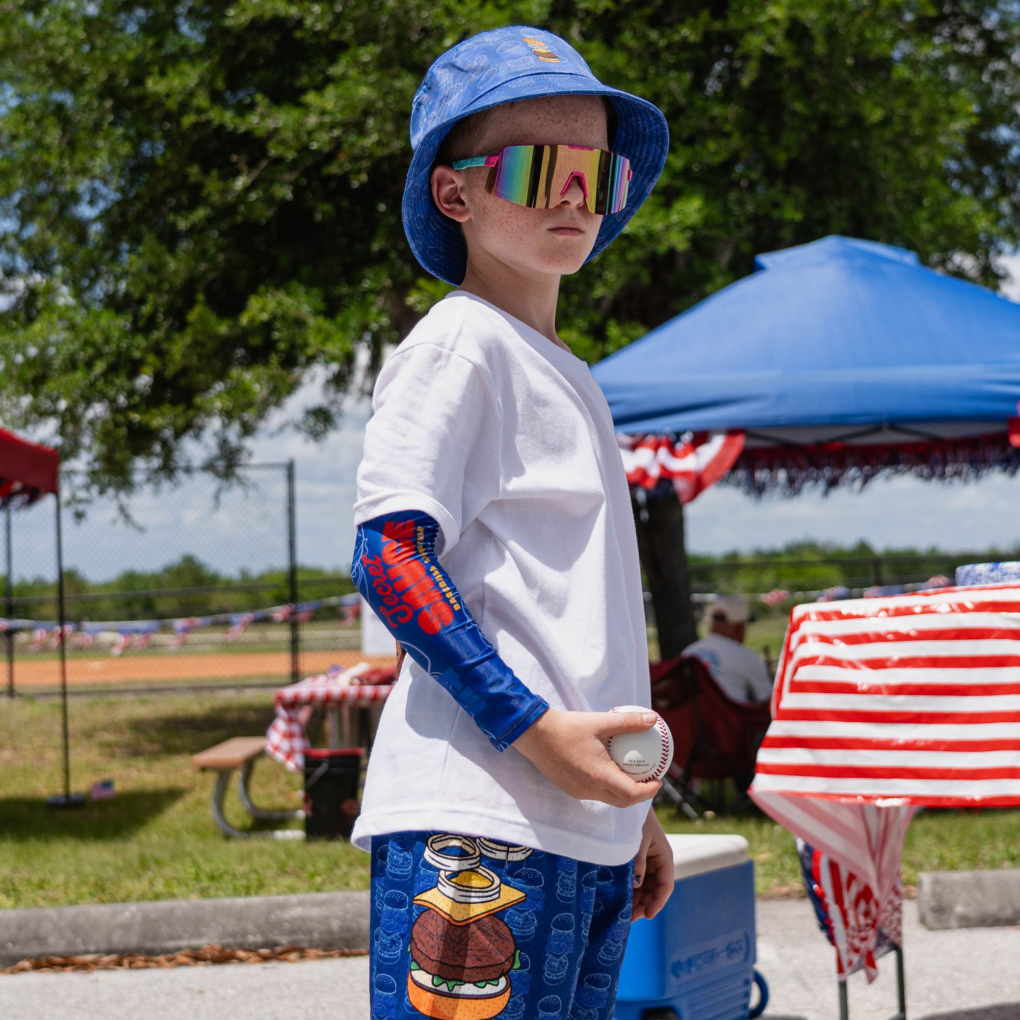 Secret Sauce Arm Sleeve - Blue: Side-facing youth male model wearing blue Secret Sauce arm sleeve with blue burger bucket hat rainbow shield sunglasses and white tee holding baseball at outdoor baseball event