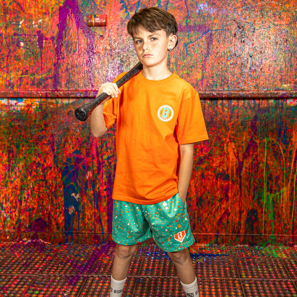 Child in orange BL101 shirt and green splatter shorts holding a bat against a colorful wall.