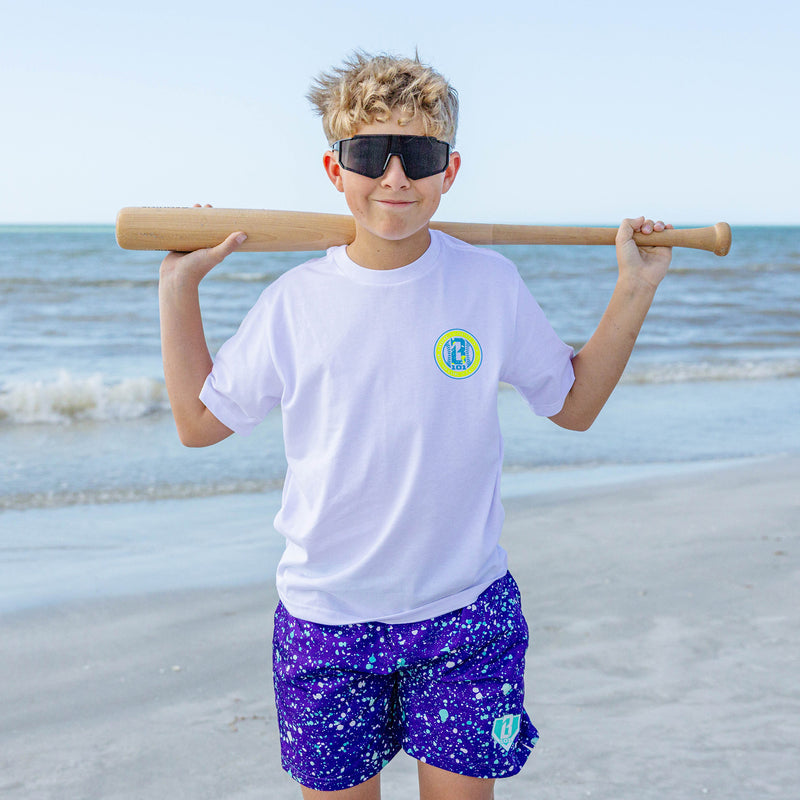 Young boy on the beach wearing BL101 youth purple and teal splatter swim shorts with a white BL101 tee, holding a wood bat across his shoulders.