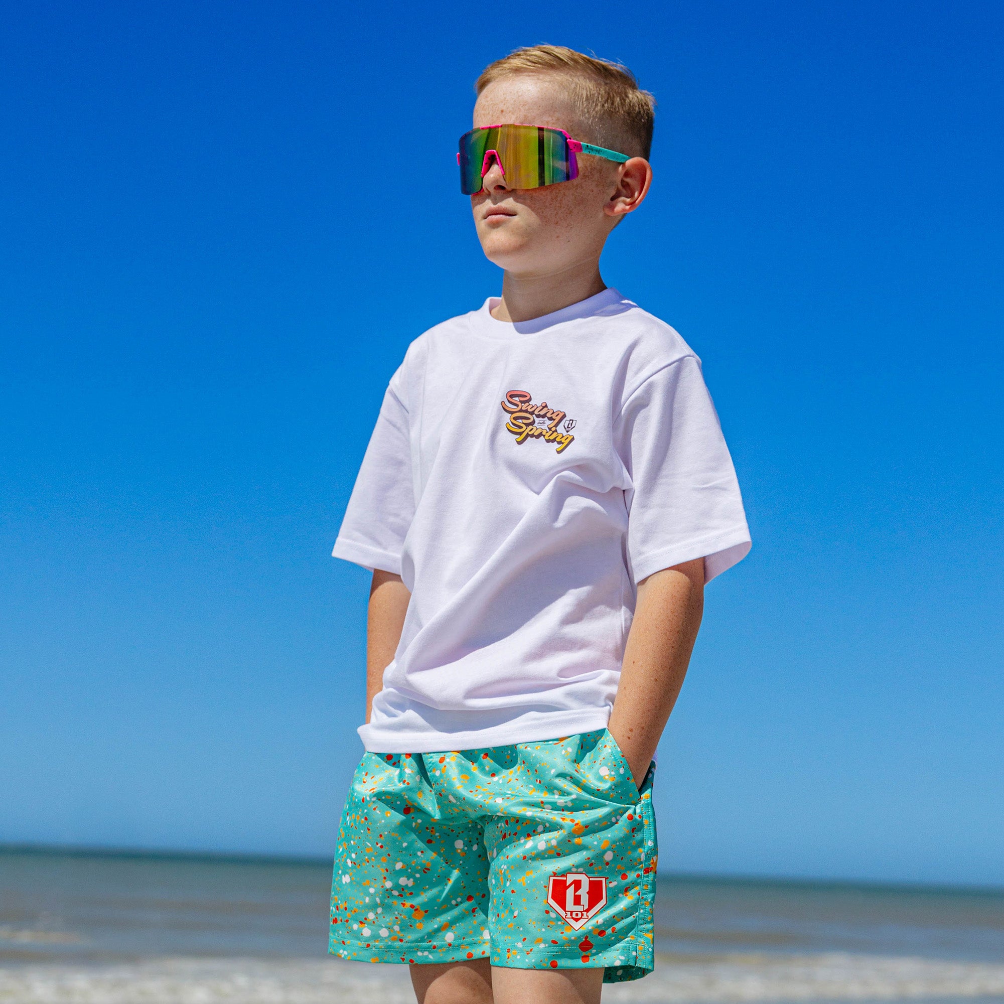 Front view of BL101 teal splatter print swim trunks worn poolside with a white tee, showing the BL101 logo on the leg.