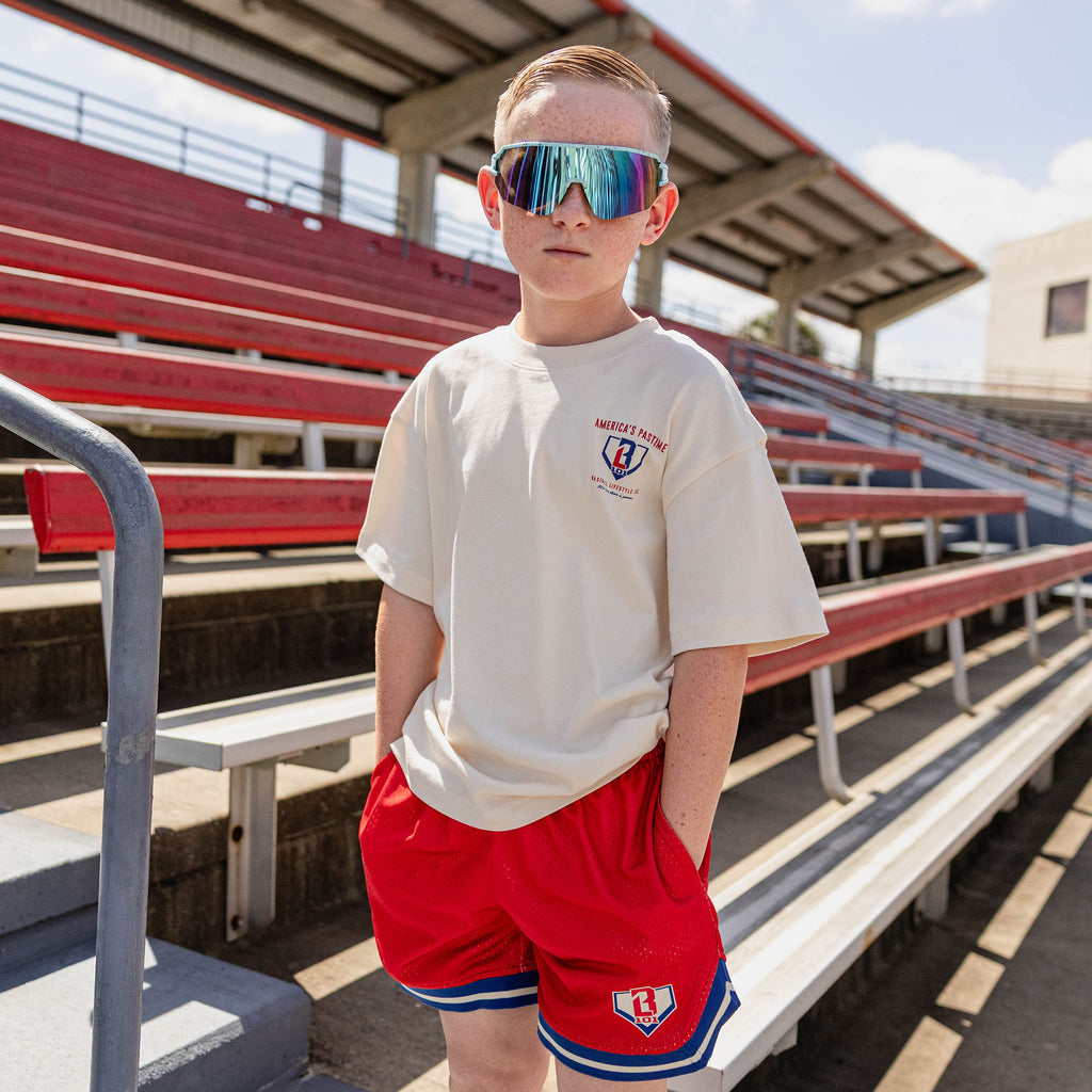Young boy wearing the BL101 youth cream tee and red mesh shorts in a stadium, wearing shield sunglasses.