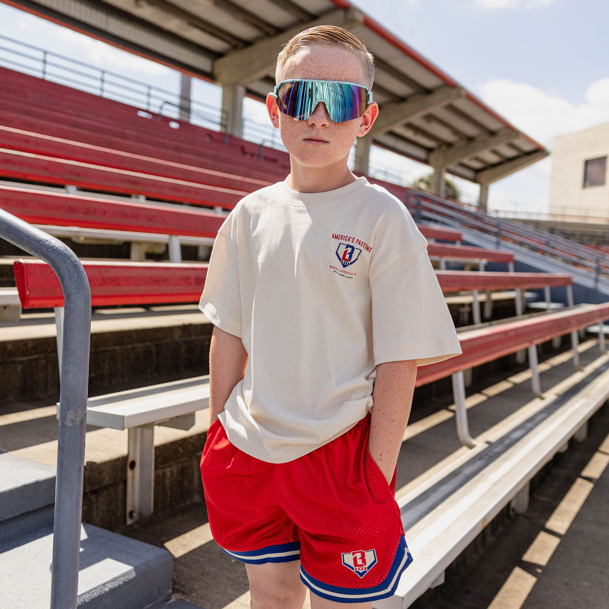 Young boy wearing the BL101 youth cream tee and red mesh shorts in a stadium, wearing shield sunglasses.