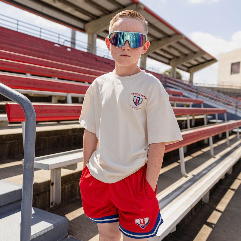 Young boy wearing the BL101 youth cream tee and red mesh shorts in a stadium, wearing shield sunglasses.