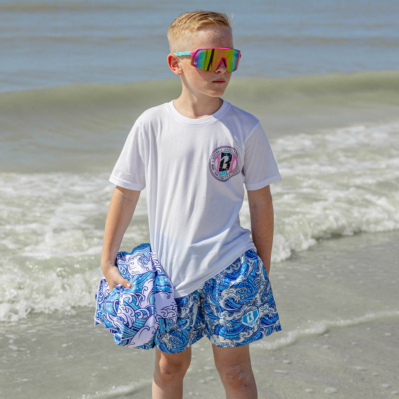 Boy standing at the shoreline wearing BL101 wave print swim trunks and a white BL101 graphic tee, holding a matching wave print towel and wearing colorful shield sunglasses.