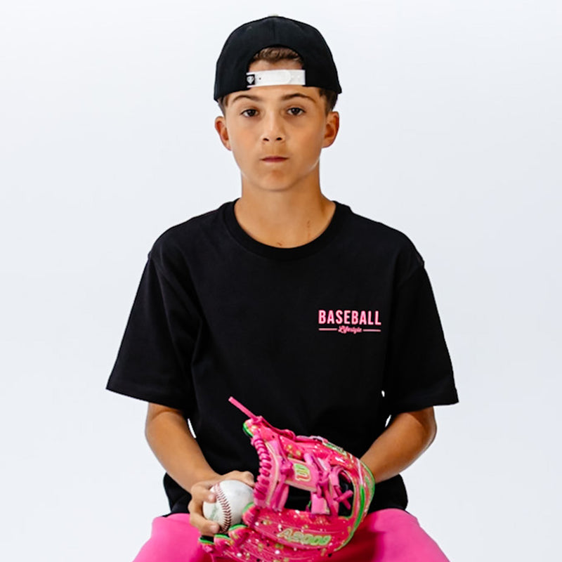 Child wearing a black t-shirt with pink 'BASEBALL Lifestyle' text in top corner, holding a pink baseball glove and ball on a white background