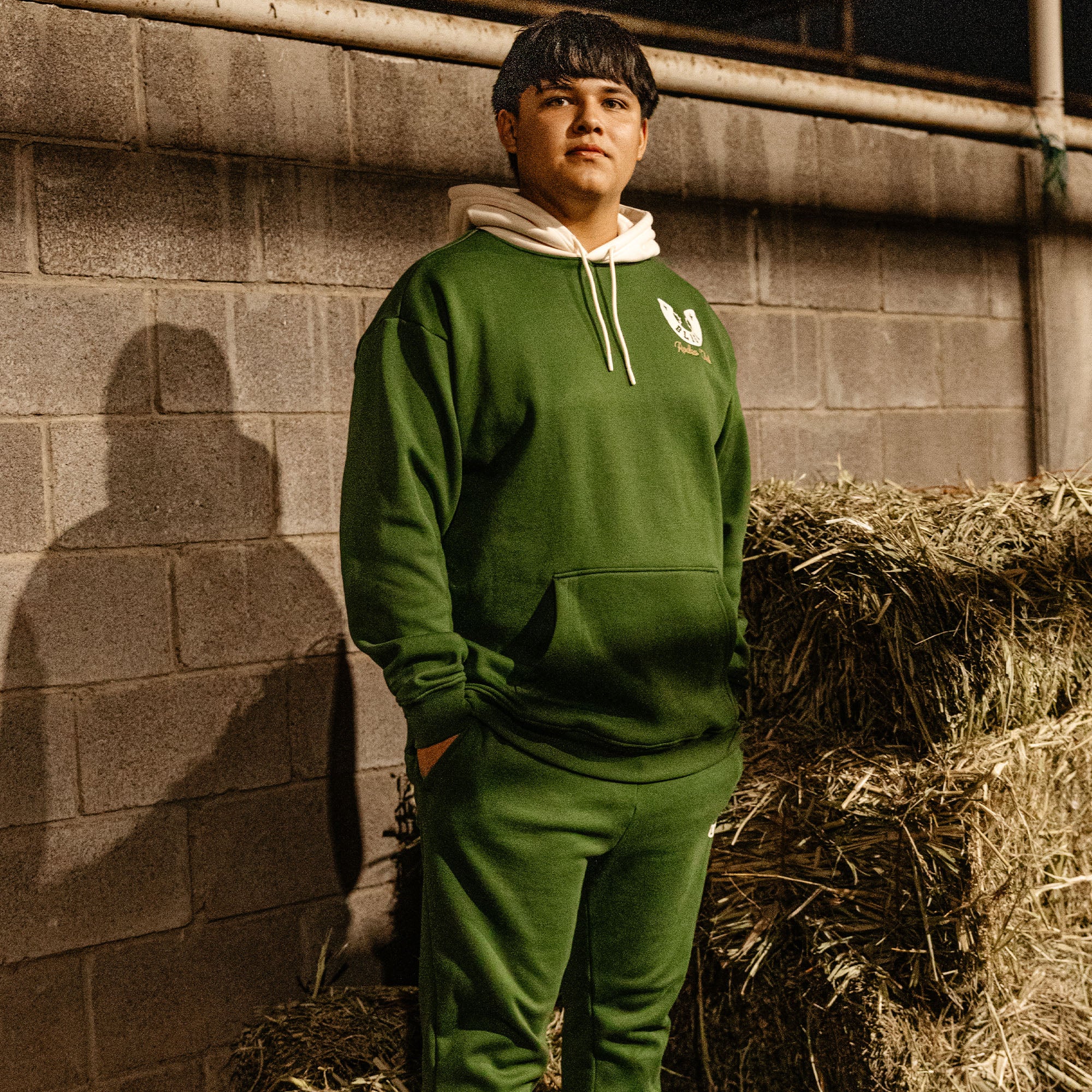 Person wearing a green and white hoodie and pants standing next to a stack of hay against a brick wall.