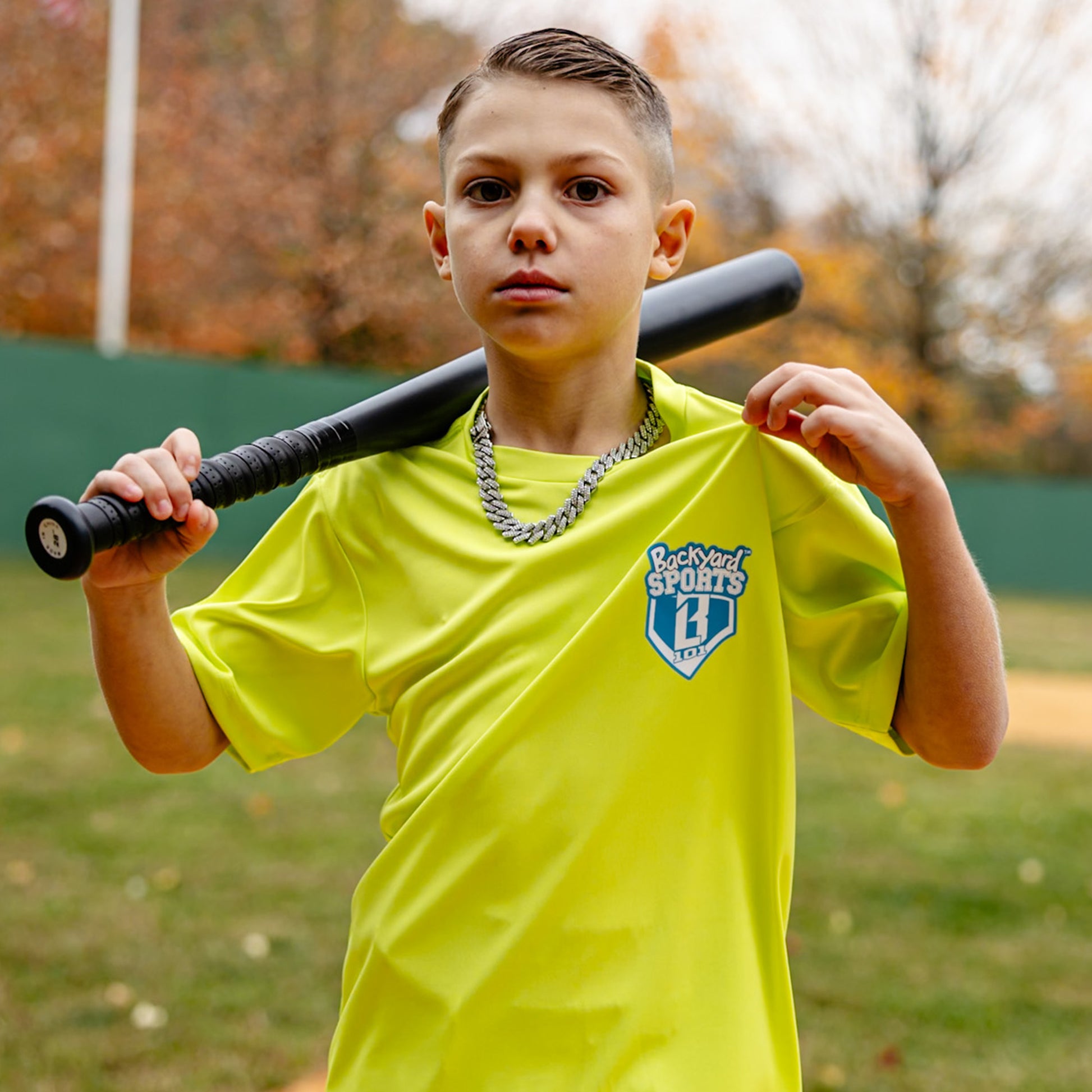 Child holding a baseball bat wearing a yellow 'Backyard Sports BL101' shirt outdoors.