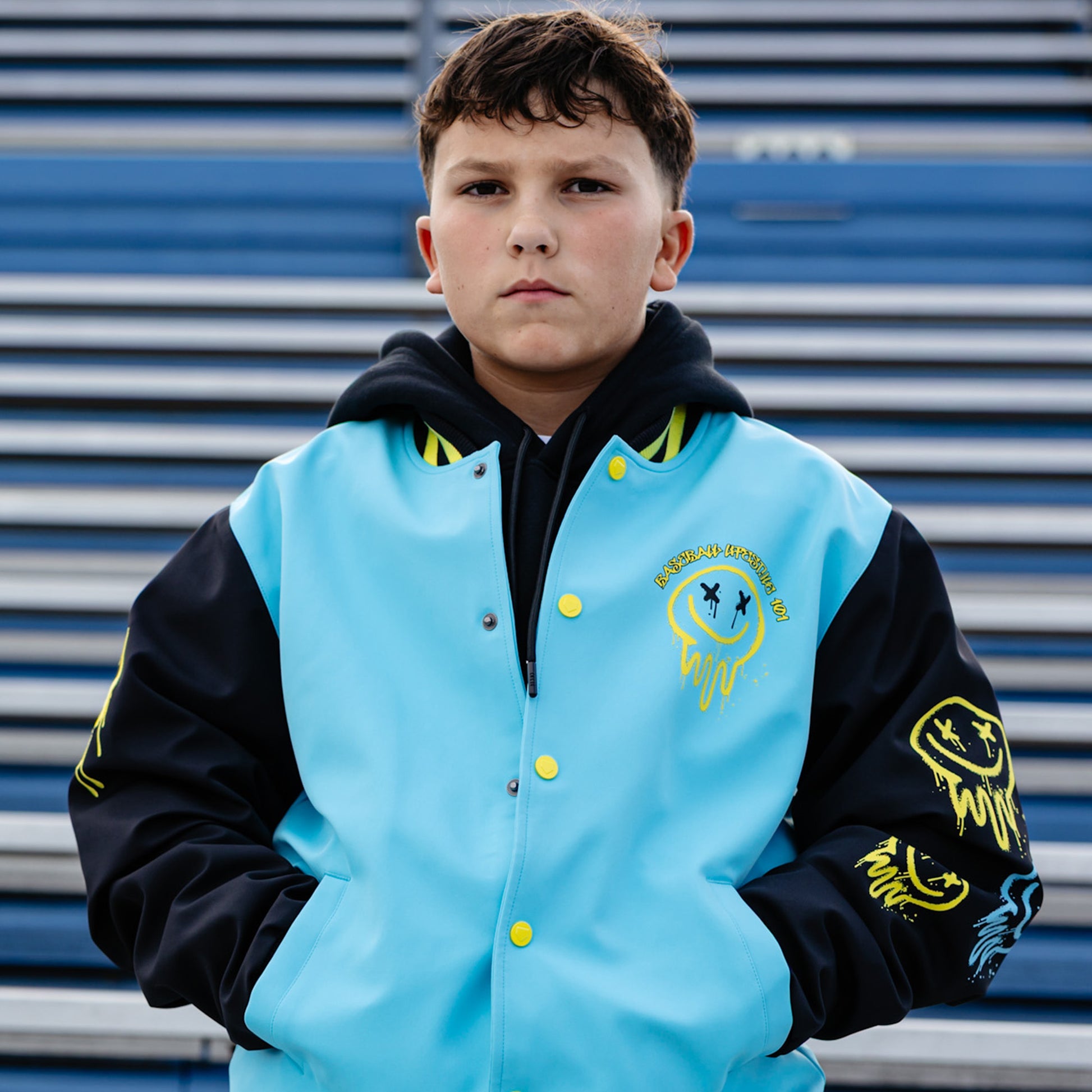 Young boy wearing a blue, yellow and black varsity jacket with a yellow smiley face, standing in front of blue stadium seats.