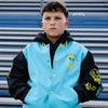 Young boy wearing a blue, yellow and black varsity jacket with a yellow smiley face, standing in front of blue stadium seats.