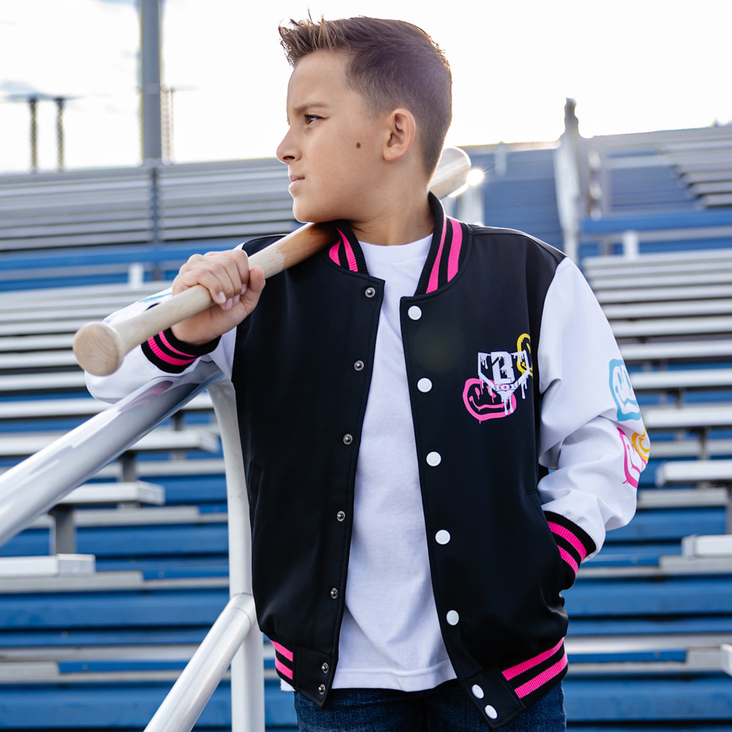Child wearing a black, pink and white varsity jacket with colorful smiley faces and white BL101 logo, holding a baseball bat in bleachers.
