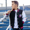 Child wearing a black, pink and white varsity jacket with colorful smiley faces and white BL101 logo, holding a baseball bat in bleachers.