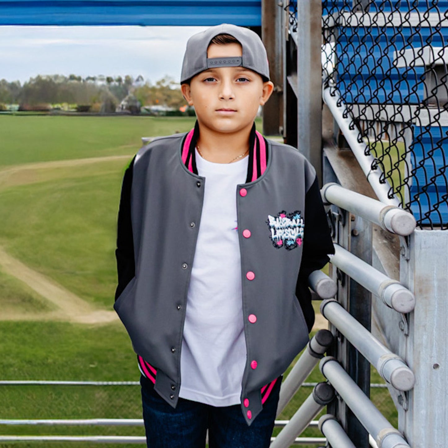 Young boy wearing a gray and black bomber jacket with pink accents, standing on bleachers at a sports field.
