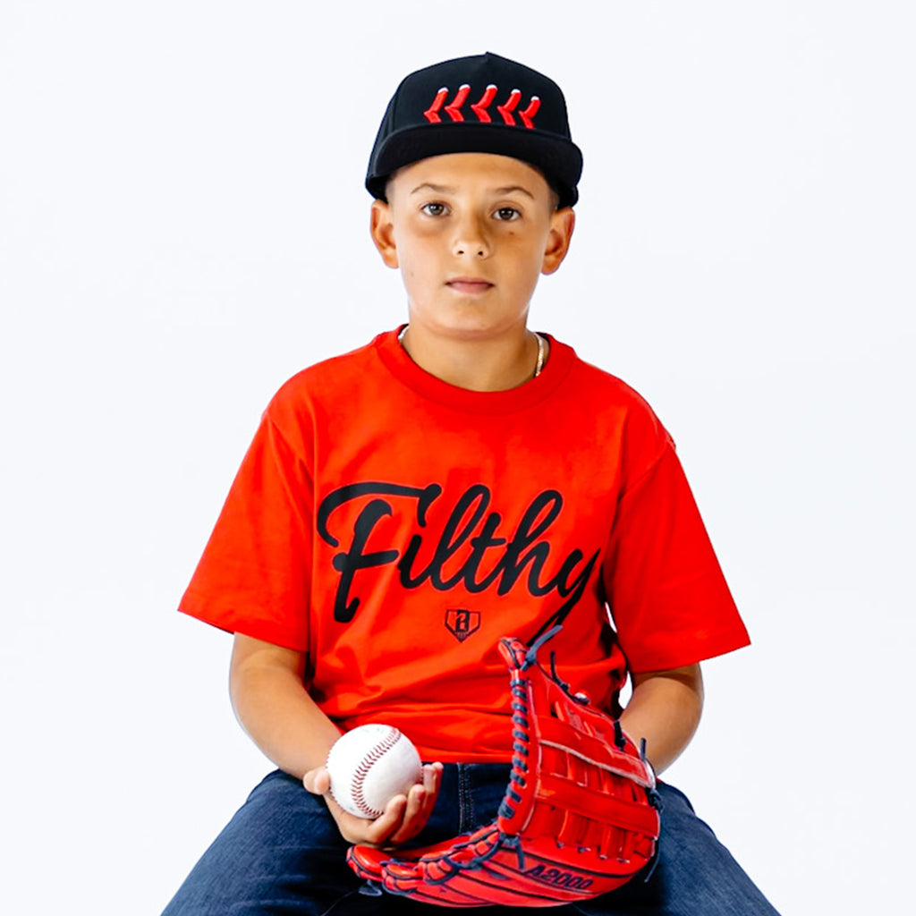 Child wearing a red 'Filthy' t-shirt, black cap, and holding a baseball and glove on a white background