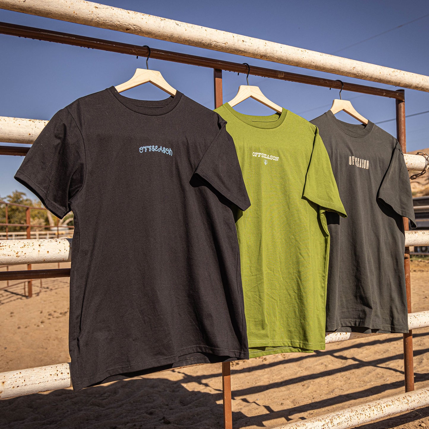 Three t-shirts in black, green, and gray with 'OFFSEASON' text hanging on a rack with a clear blue sky in the background.