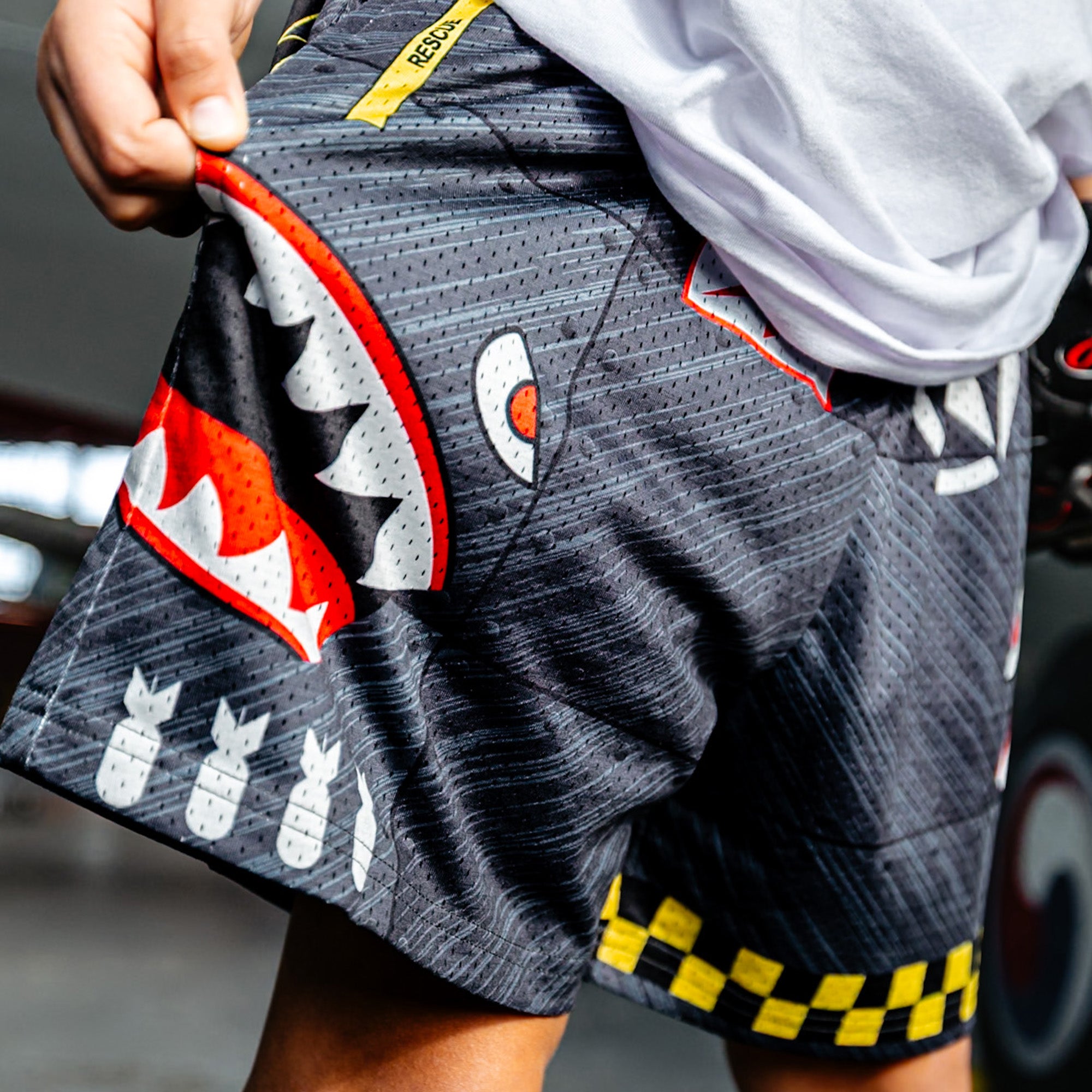 Close up of Military-inspired black athletic shorts featuring shark mouth graphics, a white star emblem, a red ‘Keep Clear’ detail, white BL101 logos on the sides, a green waistband, and yellow checkerboard trim.