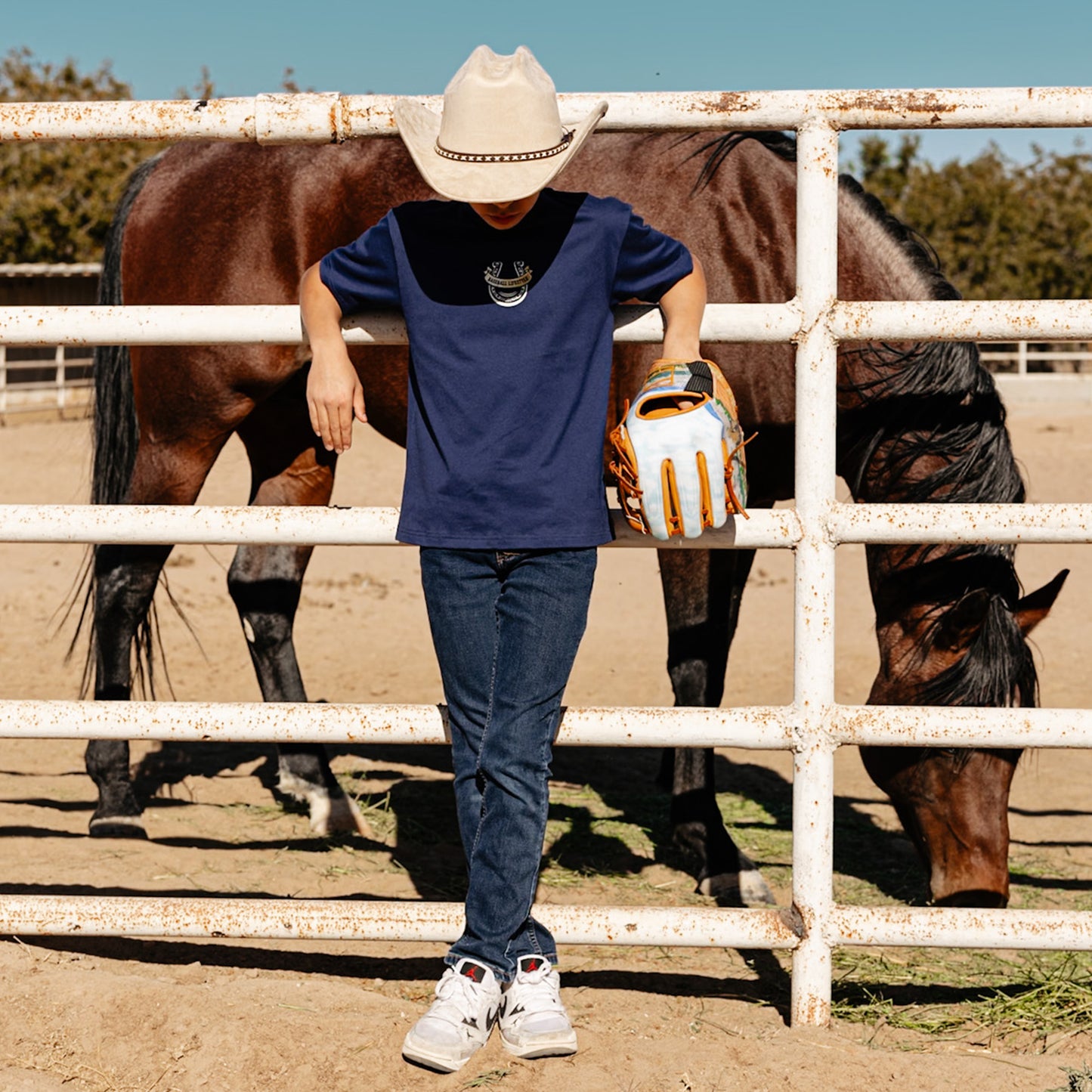 Person wearing a cowboy hat, t-shirt with a Baseball Lifestyle horseshoe logo and baseball glove standing next to a horse behind a fence.