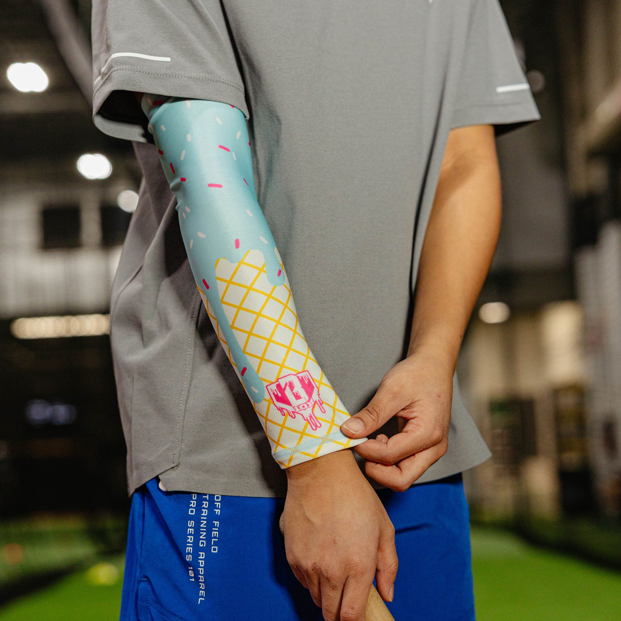 Youth player in a gray shirt and blue shorts wearing the cotton candy ice cream arm sleeve on their left arm while adjusting it at the wrist in an indoor facility