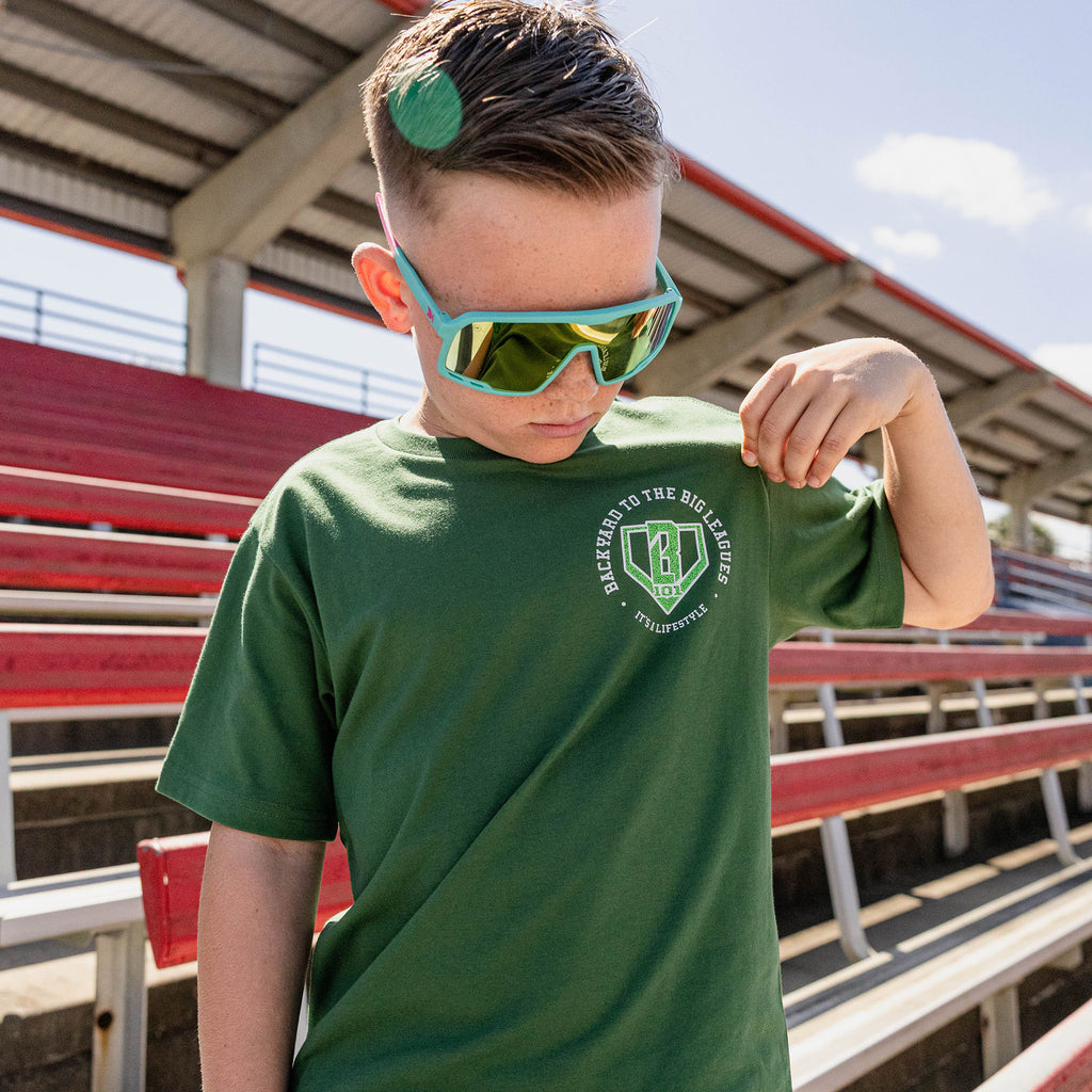 Boy looking down pulling his sleeve at red stadium bleachers wearing teal shield sunglasses and a forest green tee with a left chest BL101 logo graphic