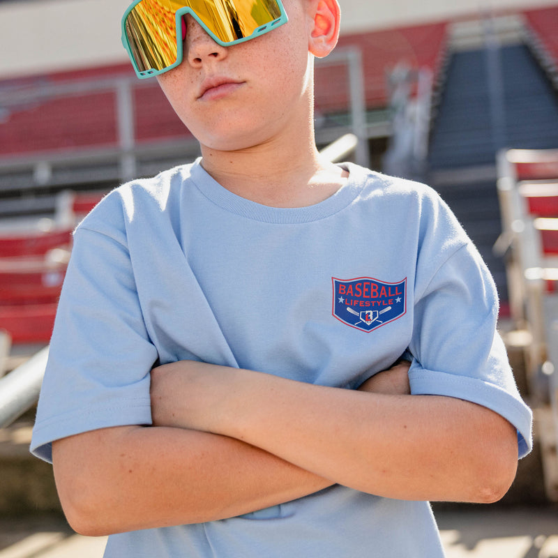 Close up of boy with arms crossed wearing teal shield sunglasses and a light blue tee with a left chest Baseball Lifestyle shield graphic