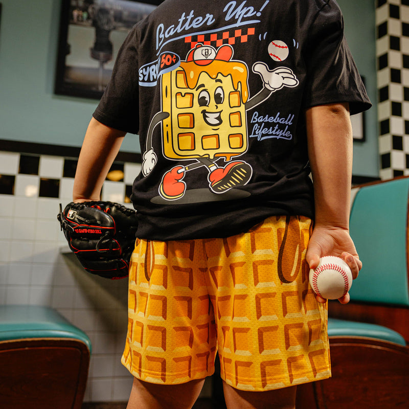 Person wearing a black t-shirt with a cartoon waffle design and waffle shorts, holding a baseball in a casual setting.