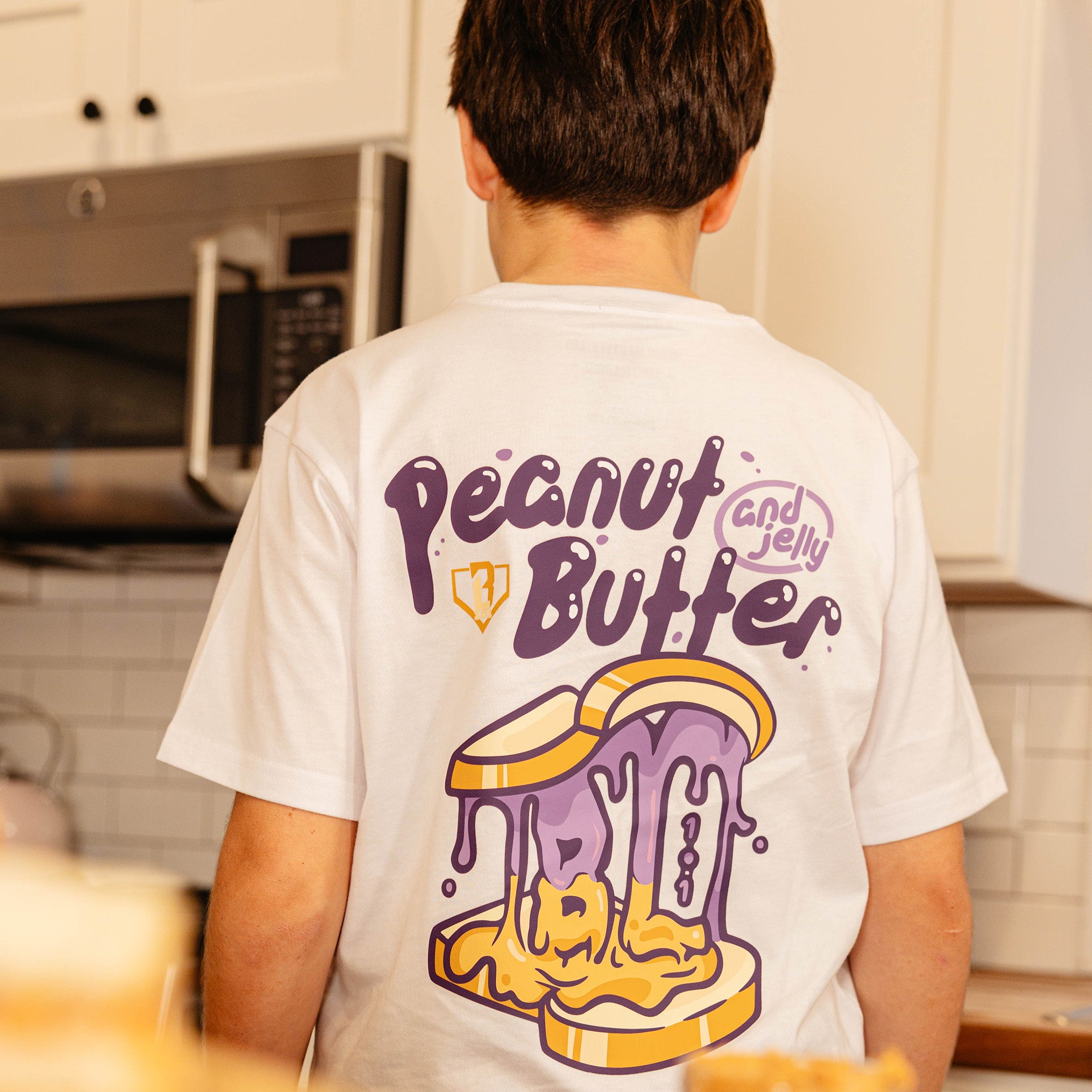 Back view of the BL101 youth Peanut Butter and Jelly white tee on a young boy in a kitchen, showing the dripping sandwich graphic.