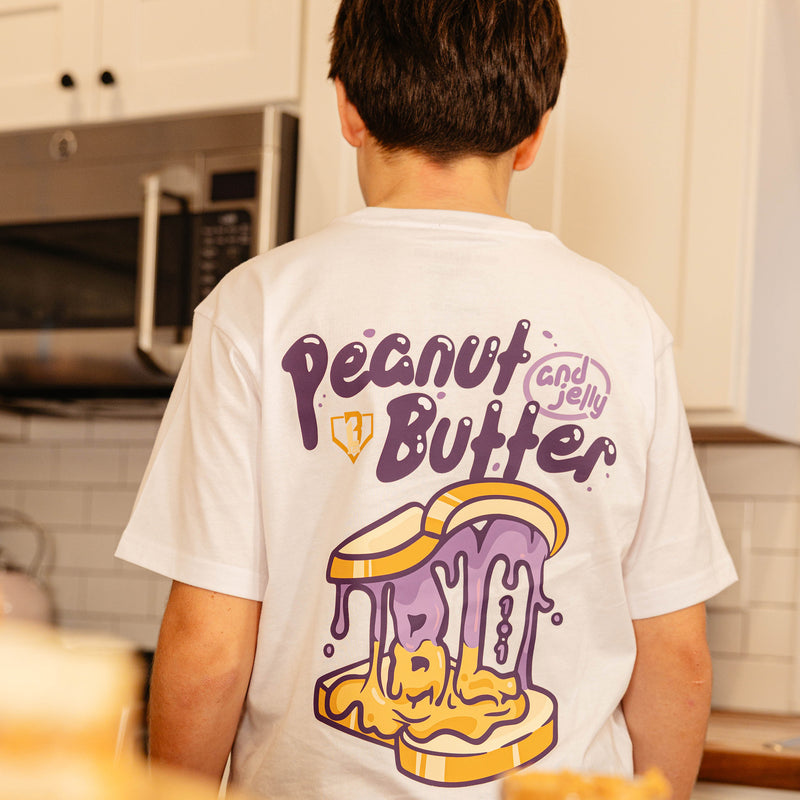 Back view of the BL101 youth Peanut Butter and Jelly white tee on a young boy in a kitchen, showing the dripping sandwich graphic.