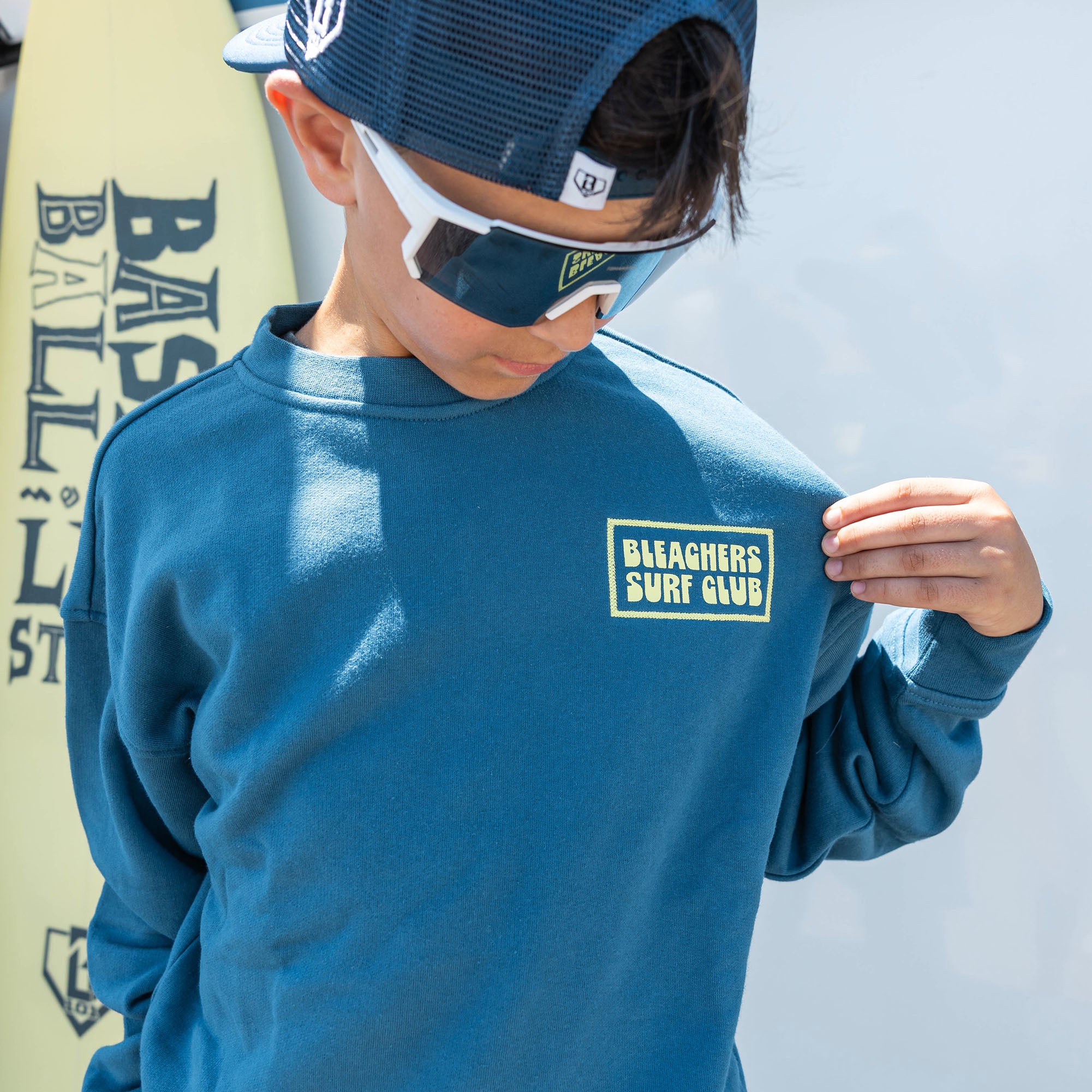Youth model wearing blue youth crewneck with "Bleachers Surf Club" design