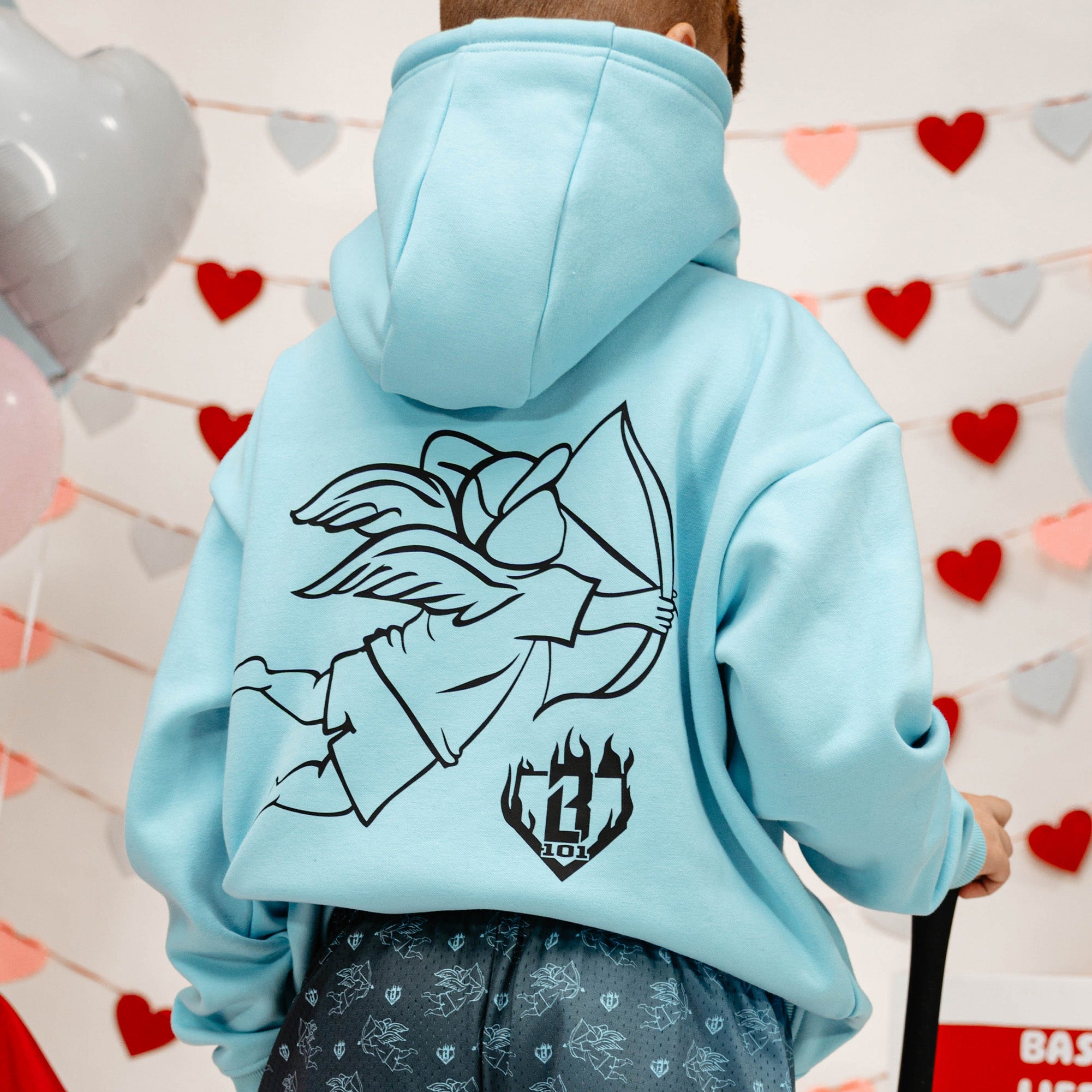 Light blue hoodie with a boy with arrow graphic design on the back, worn by a person, against a heart-patterned wall.