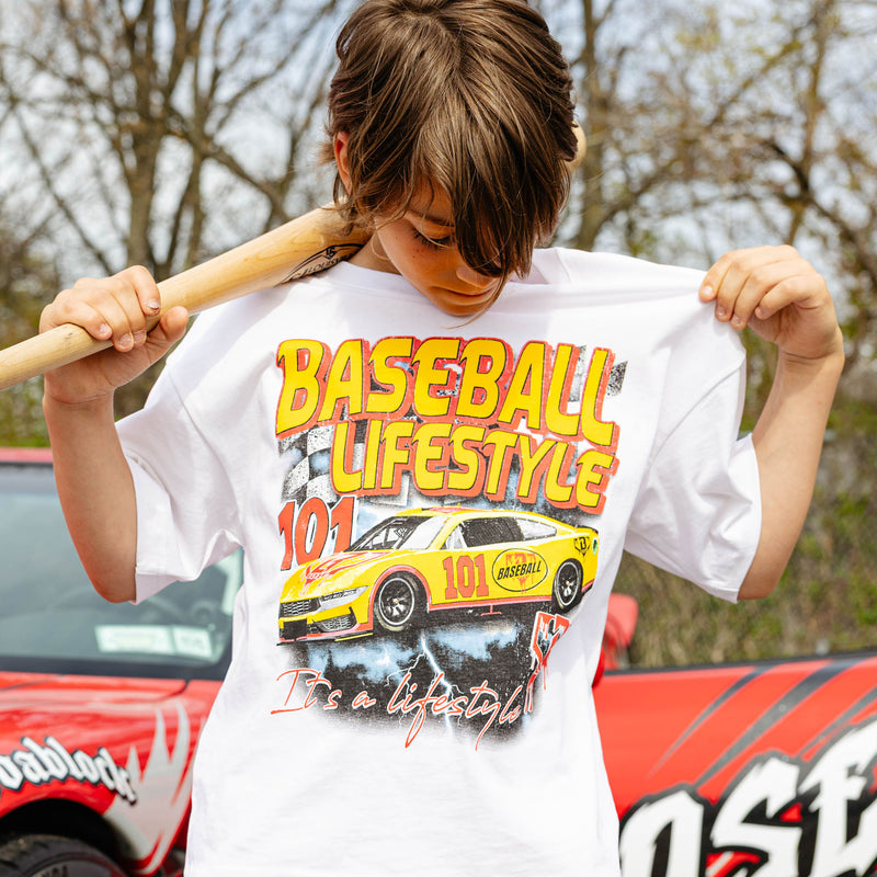 Young boy holding wooden bat across shoulders, wearing white Diamond Drift tee with yellow and red BL101 race car graphic and red flame shorts, in front of wrapped red car.