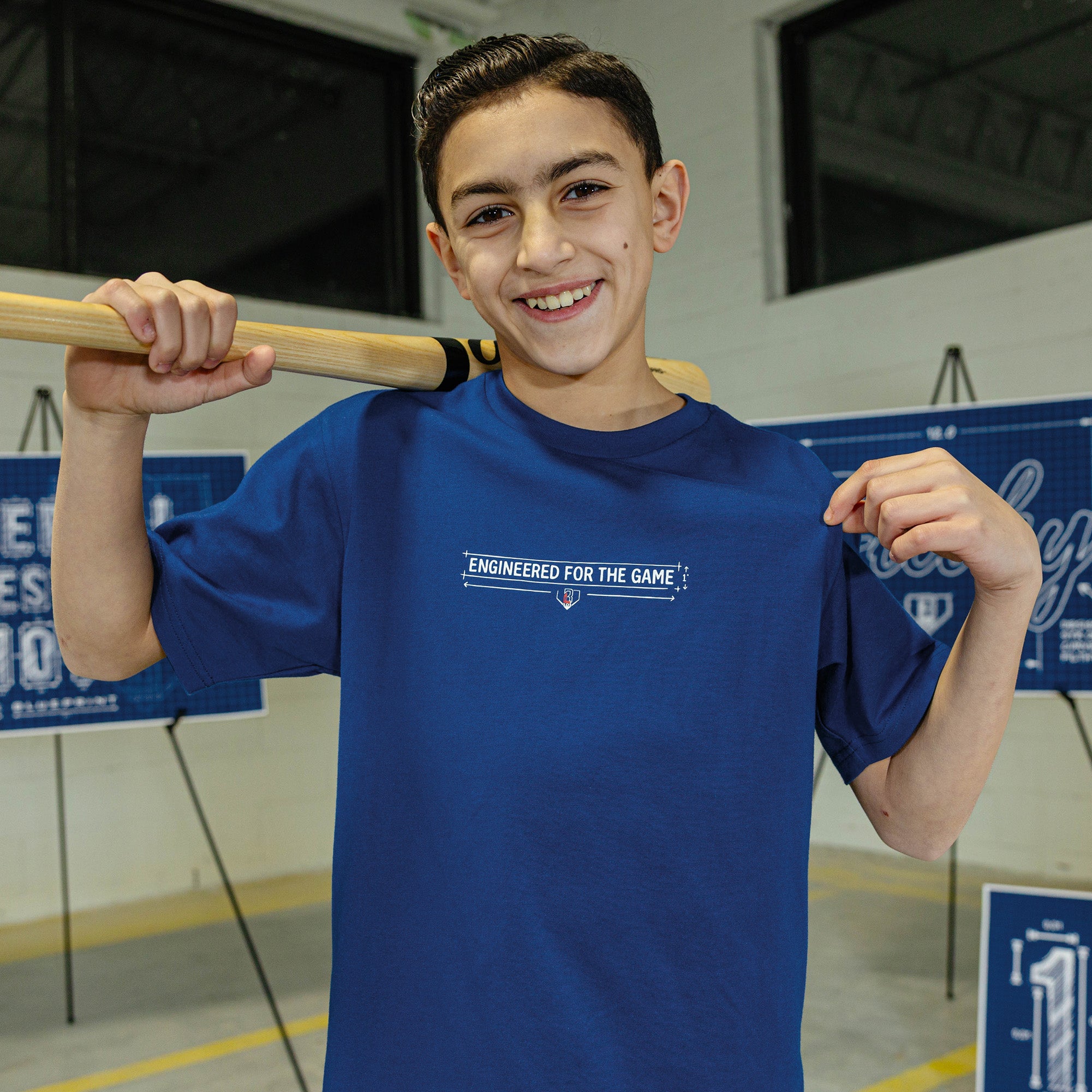Young boy wearing a blue t-shirt with a BL101 logo and "Engineered for the game" text, holding a baseball bat indoors.