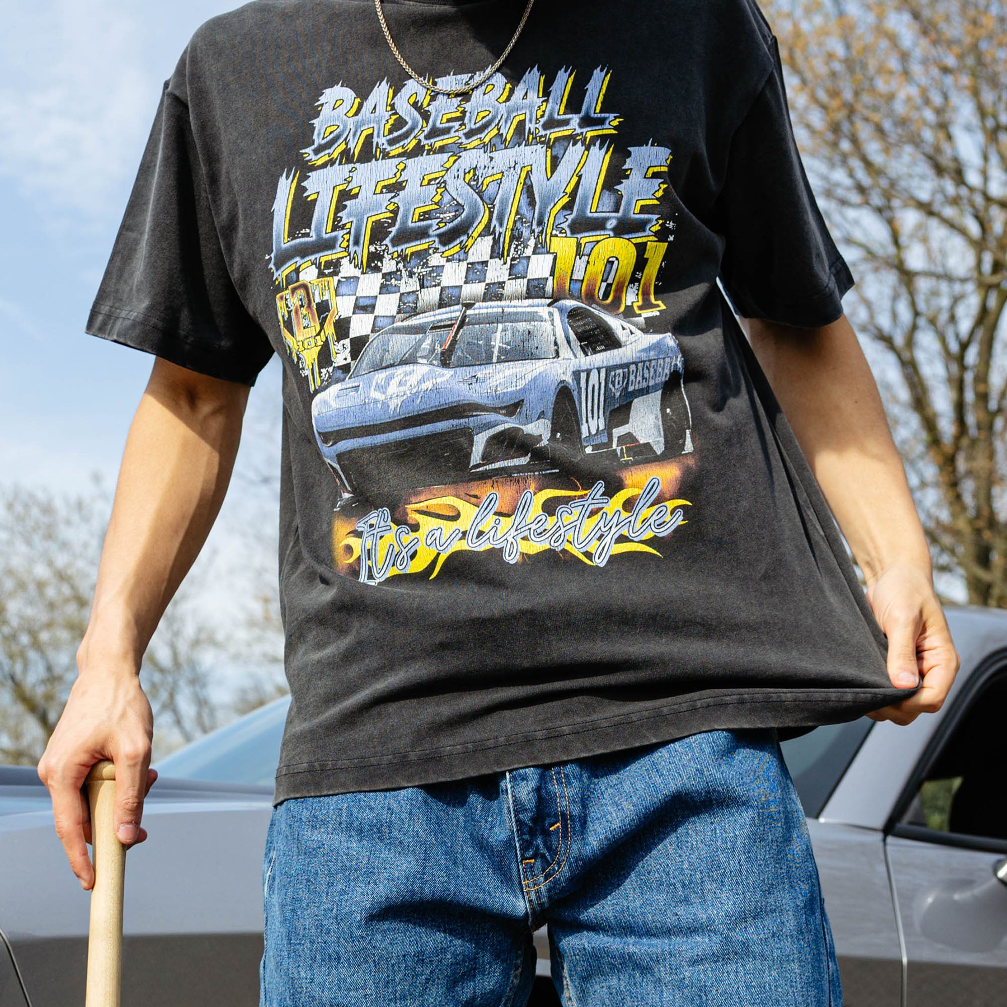 Cropped front view of washed black youth tee with full race car and flame graphic, model holding baseball bat beside silver car.