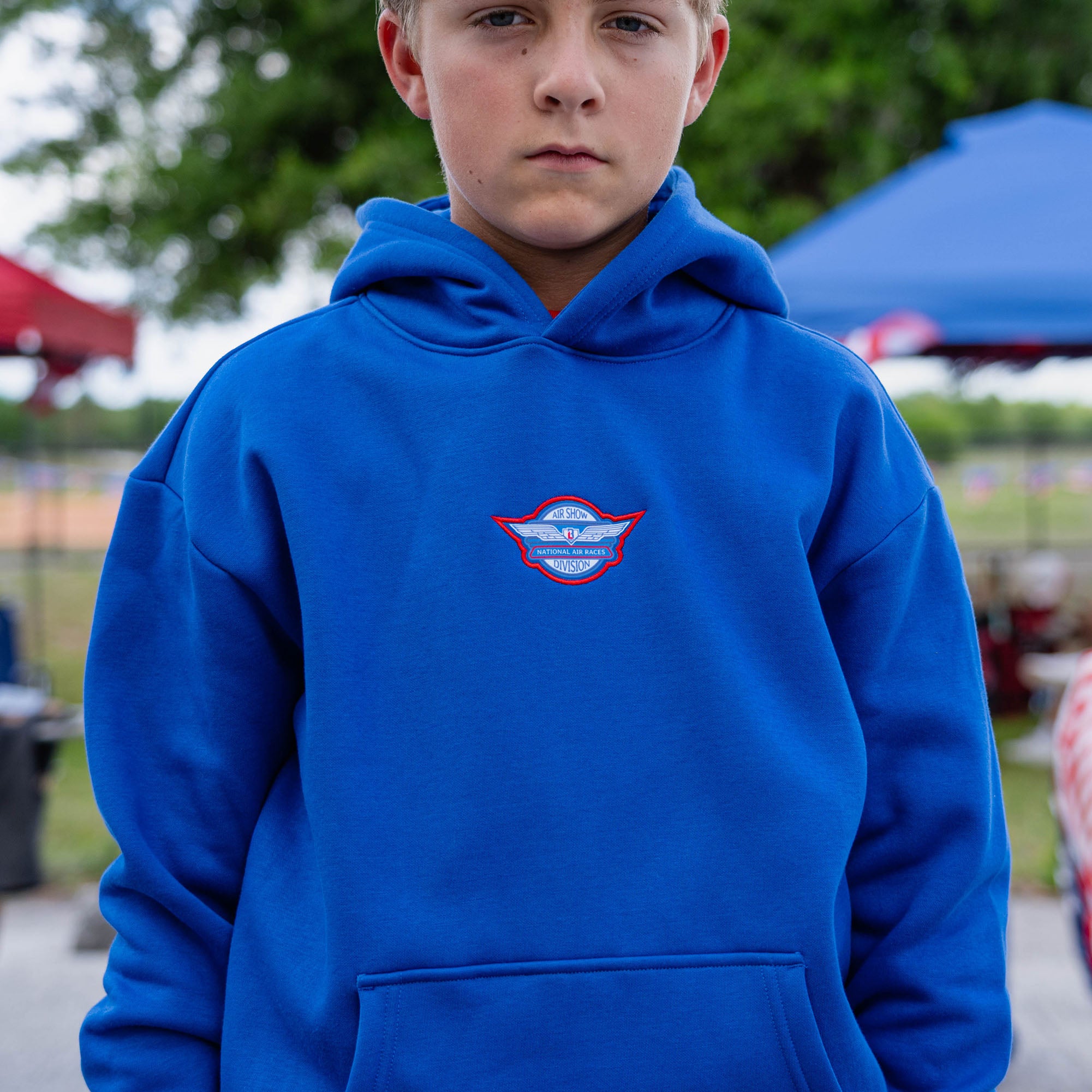 Close-up of youth male model in royal blue Fastball Flight hoodie showing Air Show Division badge on center chest at outdoor event