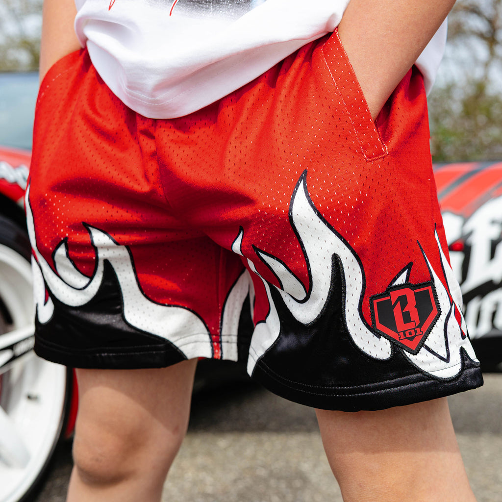 Close-up of red youth mesh shorts with black and white flame hem graphic and BL101 diamond logo, beside red car.