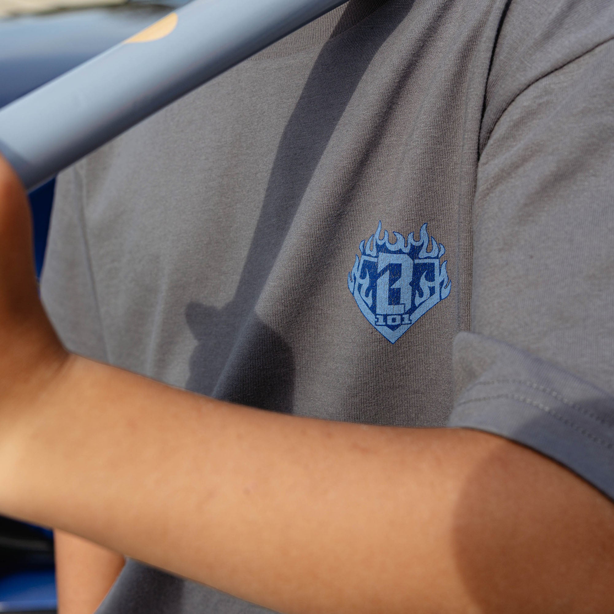Close-up of blue BL101 flame diamond chest graphic on gray youth Flame Thrower tee, baseball bat casting shadow across chest.