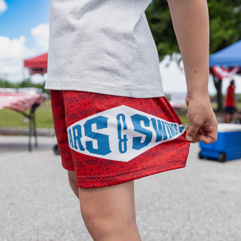 Close-up of youth model pulling fabric to show large Stars and Swings diamond logo on red shorts at outdoor event