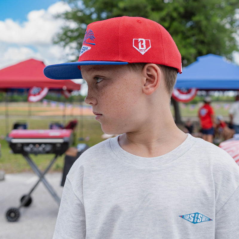 Young boy wearing a red and blue cap with a gray shirt, standing outdoors with tents and people in the background.