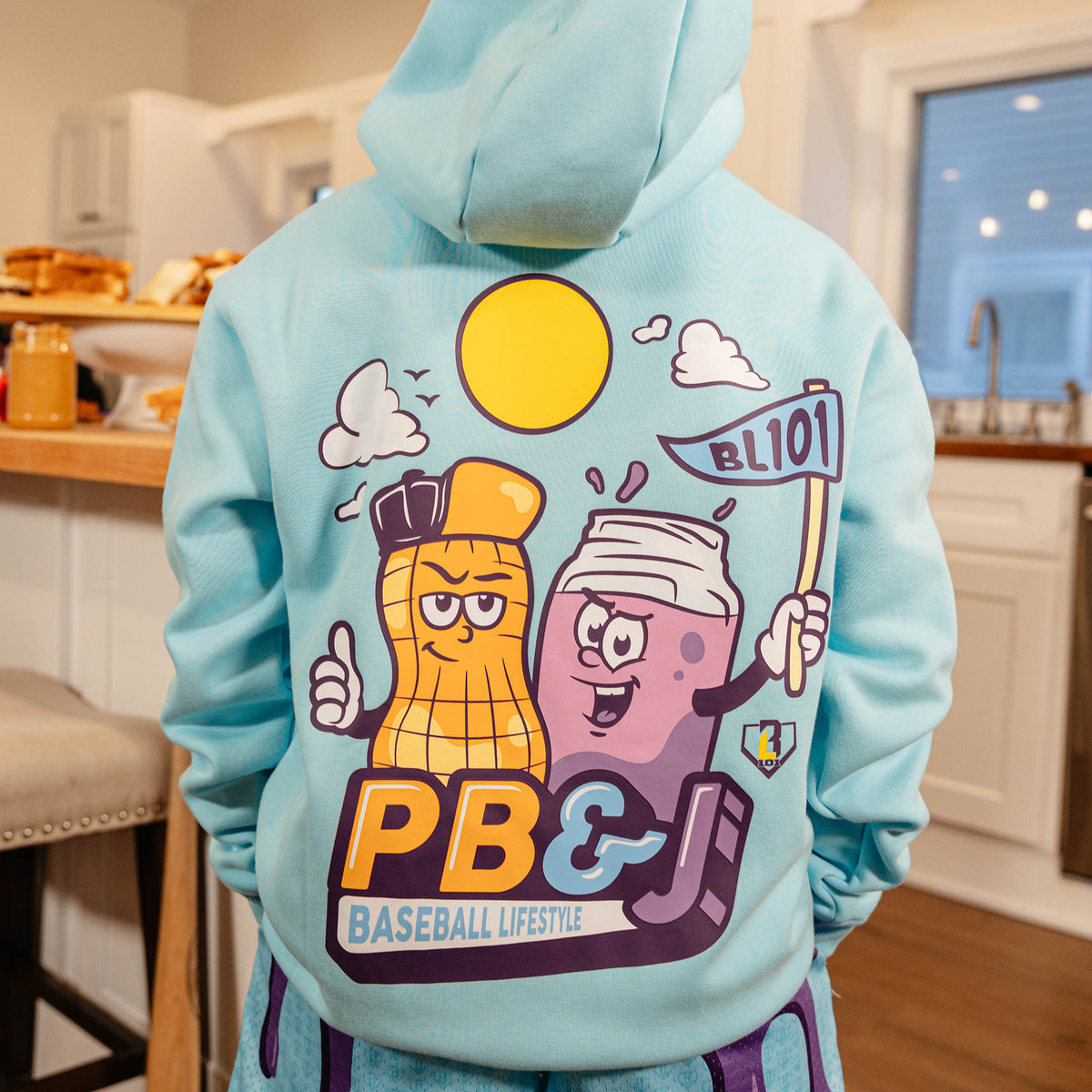 Back view of the BL101 youth PB&J Baseball Lifestyle light blue hoodie on a young boy in a kitchen, showing the cartoon peanut and jelly jar mascot graphic.