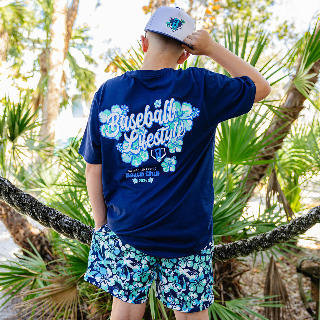 Person wearing a blue 'Baseball Lifestyle' shirt with floral designs and floral shorts in a tropical setting.
