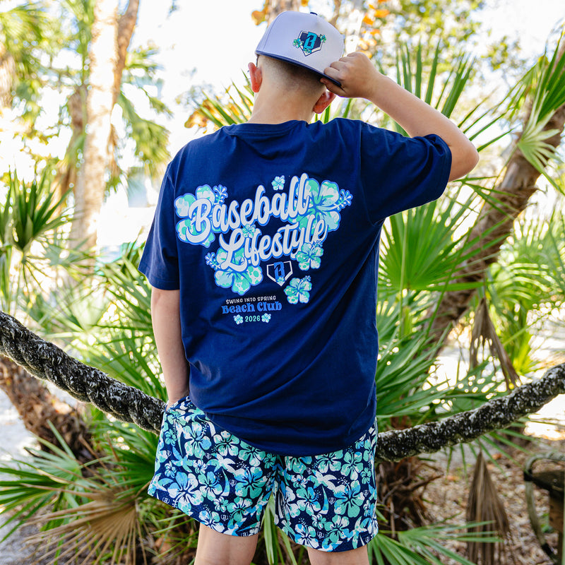 Person wearing a blue 'Baseball Lifestyle' shirt with floral designs and floral shorts in a tropical setting.