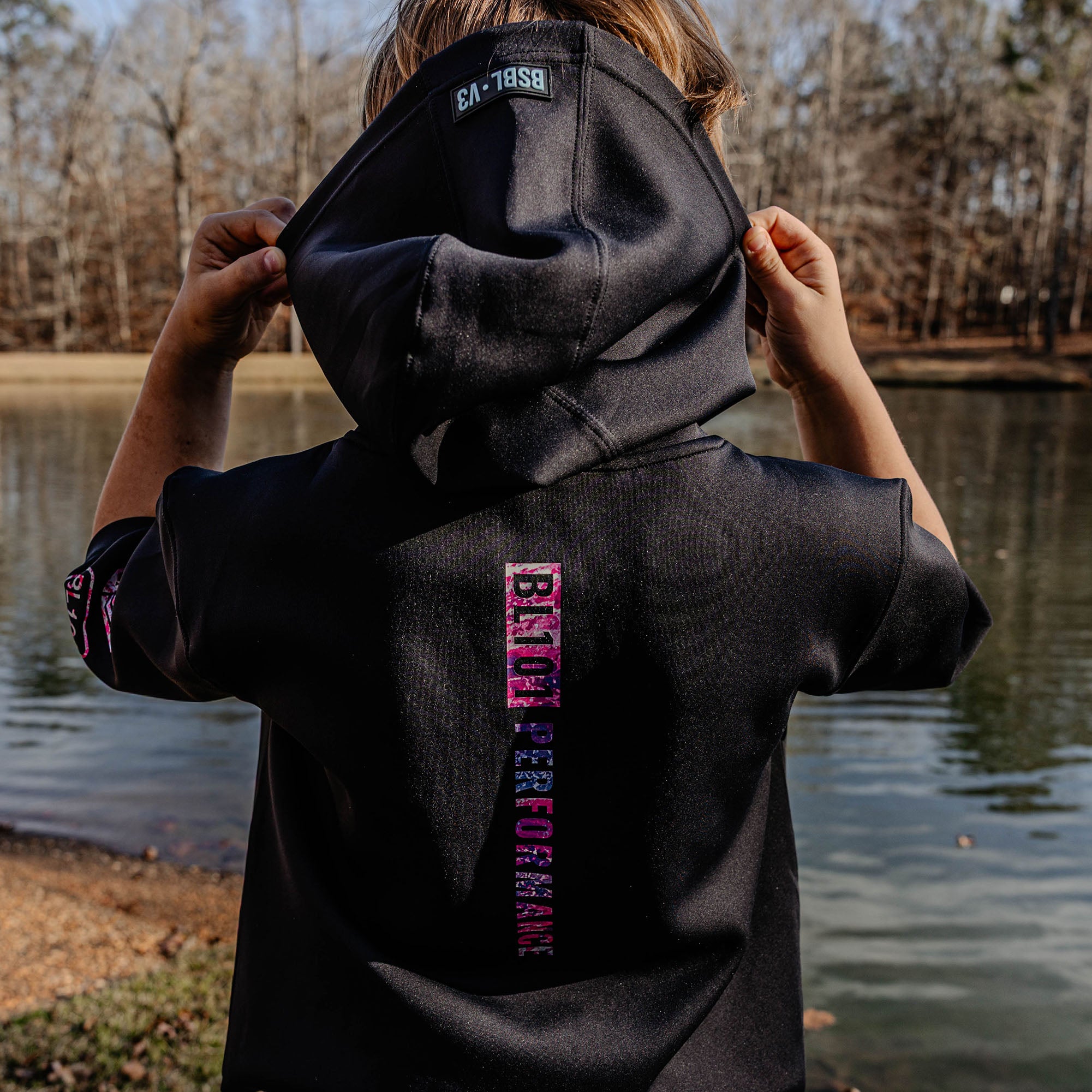 Person wearing a black hoodie with'BL101 PERFORMANCE' logo, standing by a lake.