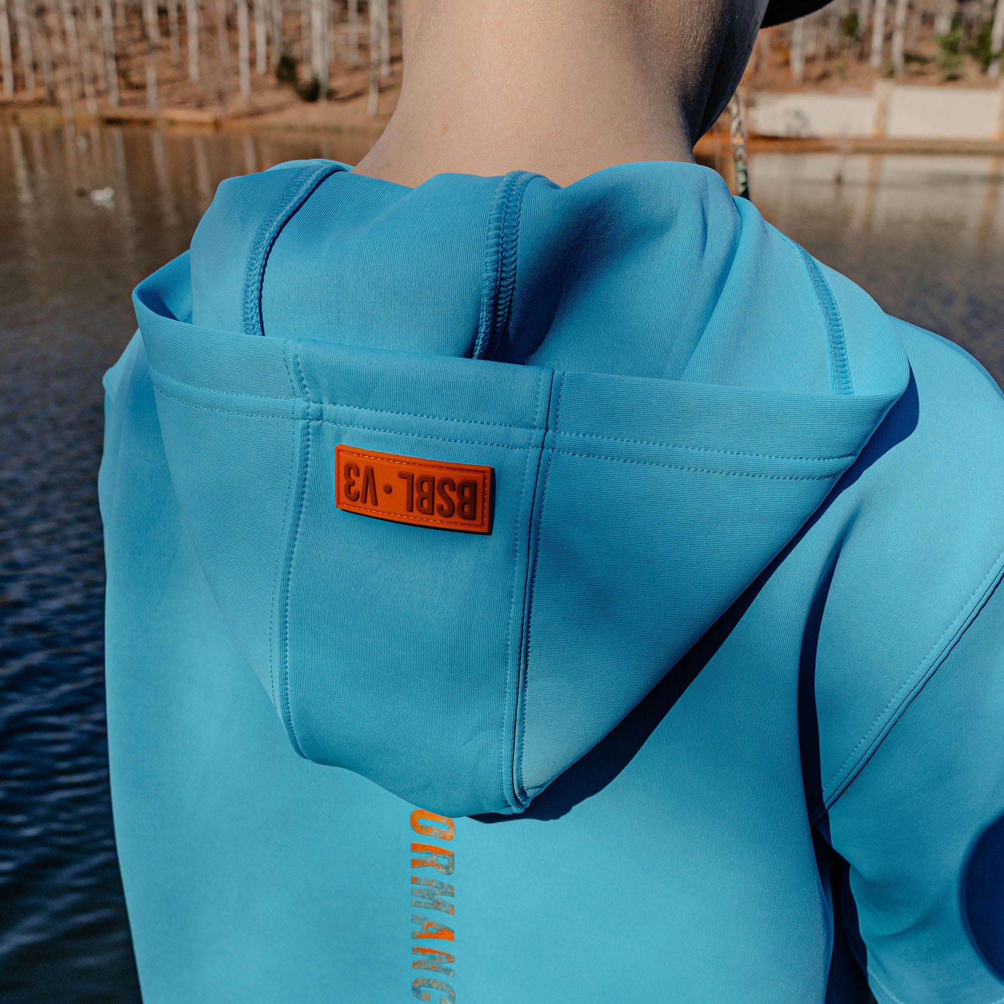 Person wearing a blue hoodie with a BSBL V3' logo, standing by a body of water.