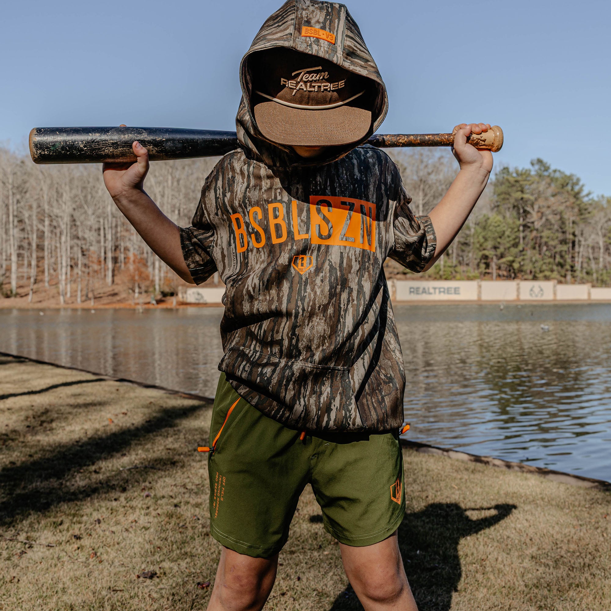 Child in camouflage outfit with orange 'BSBL SZN' text holding a baseball bat by a lake
