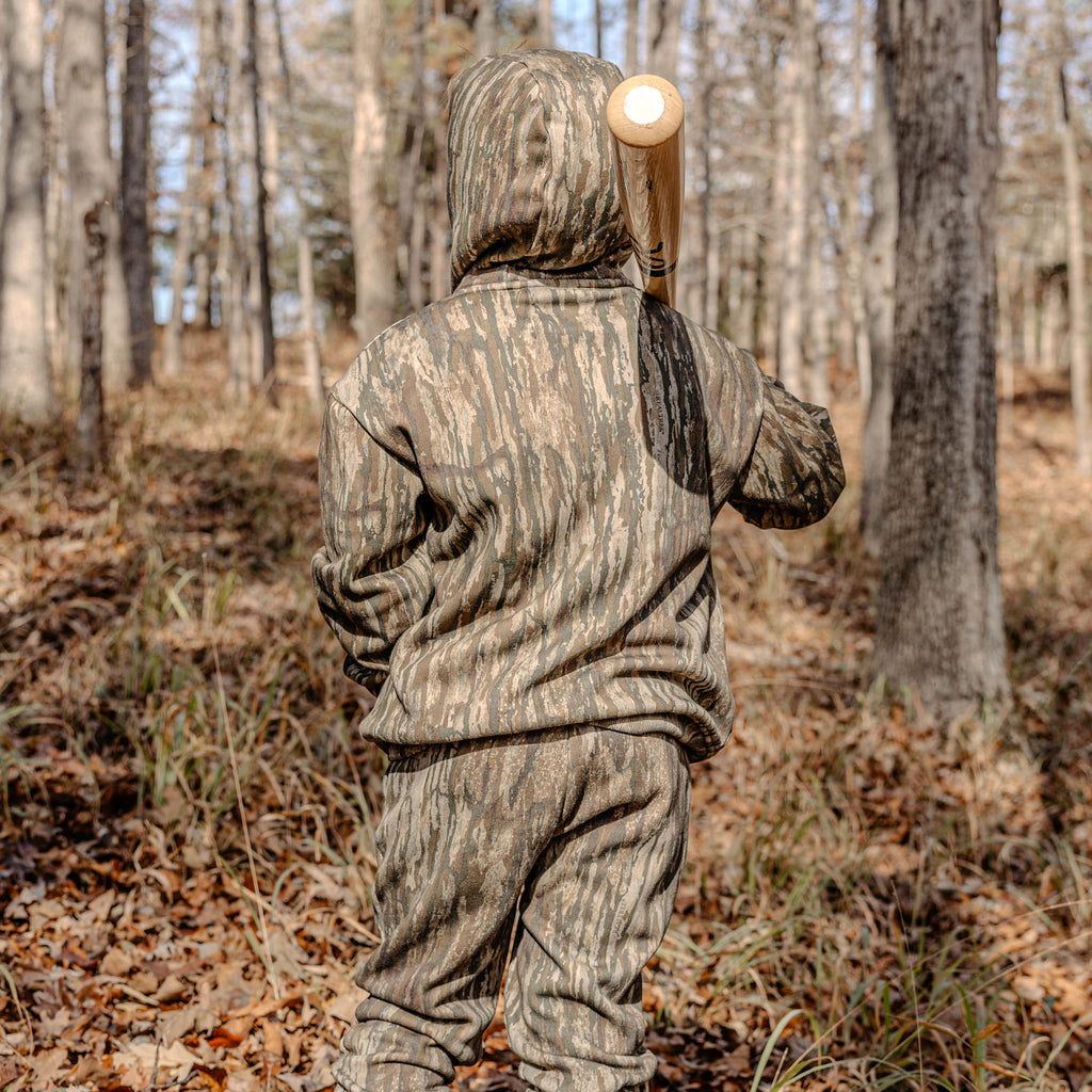 Person in camouflage hoodie and sweatpants holding a bat in a forest setting