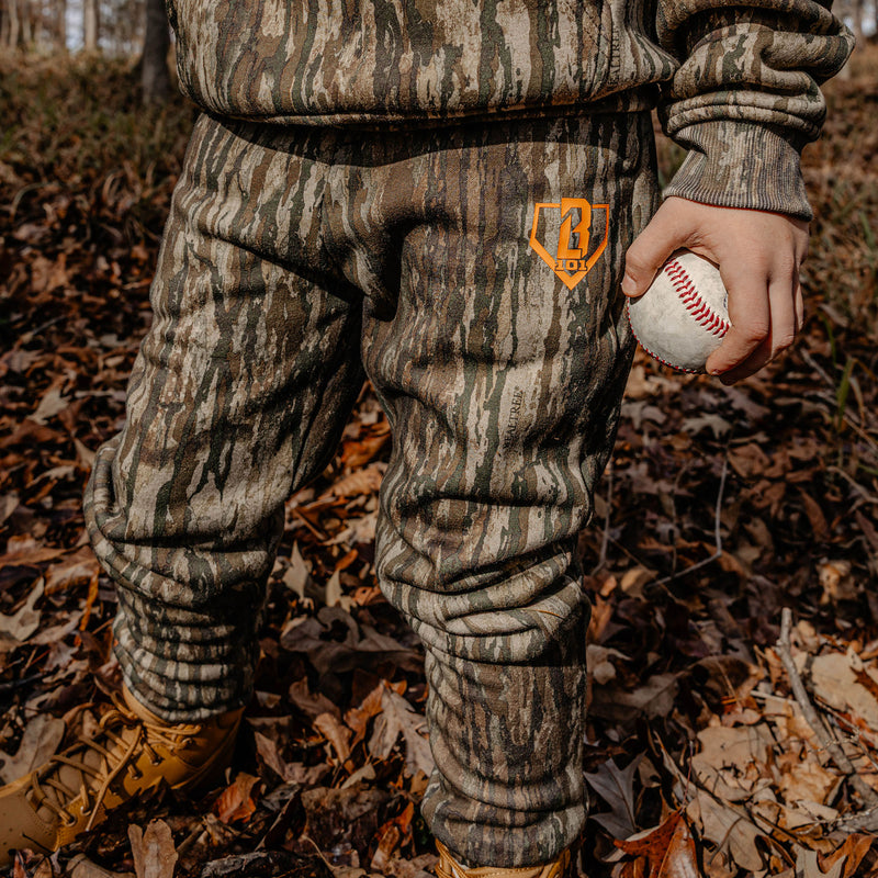 Person wearing camouflage pants with a orange BL101 logo, holding a baseball in a forest setting.